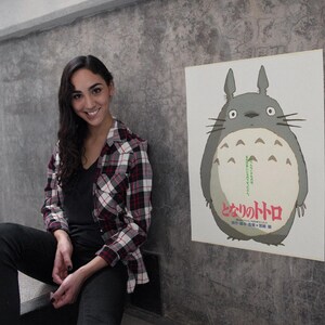 Pode incluir: Um cartaz com o personagem Totoro do filme de anima&ccedil;&atilde;o Meu Vizinho Totoro. O cartaz mostra um Totoro cinzento com uma barriga branca e texto japon&ecirc;s. O cartaz est&aacute; montado numa parede cinzenta.