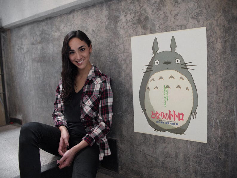 Pode incluir: Um cartaz com o personagem Totoro do filme de anima&ccedil;&atilde;o Meu Vizinho Totoro. O cartaz mostra um Totoro cinzento com uma barriga branca e texto japon&ecirc;s. O cartaz est&aacute; montado numa parede cinzenta.