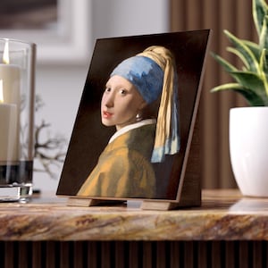 May include: A framed print of Johannes Vermeer's "Girl with a Pearl Earring" on a wooden easel. The painting features a young woman with a blue and yellow headscarf and a pearl earring. The print is displayed on a marble-like surface.