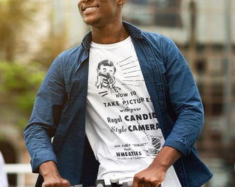 May include: A white t-shirt with the text "HOW TO TAKE PICTURES with your Regal CANDID Style CAMERA" is worn with a blue denim shirt and jeans. The person is riding a black scooter.