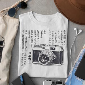 Puede incluir: Camiseta blanca con una ilustración en blanco y negro de una cámara vintage, con texto japonés. La imagen también muestra una bolsa de mano, gafas de sol y una cámara pequeña, sugiriendo un tema de fotografía.