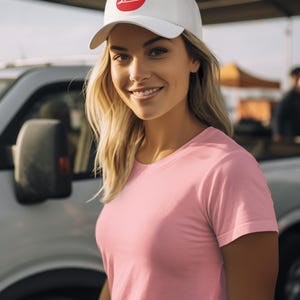 May include: A white baseball cap with a red circular logo that says "Leica". The person is wearing a pink t-shirt. The background includes a vehicle and a sunny outdoor setting.