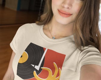 Puede incluir: Camiseta color crema con un diseño gráfico. El diseño presenta un disco de vinilo negro con centro amarillo, una mano amarilla y un fondo rojo. El gráfico está en la parte delantera de la camiseta.