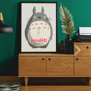 Pode incluir: Um poster emoldurado com o personagem Totoro do filme de anima&ccedil;&atilde;o Meu Vizinho Totoro. O poster &eacute; exibido em um arm&aacute;rio de madeira com uma m&aacute;quina de escrever preta, uma folha de ouro e uma lumin&aacute;ria pendente preta.