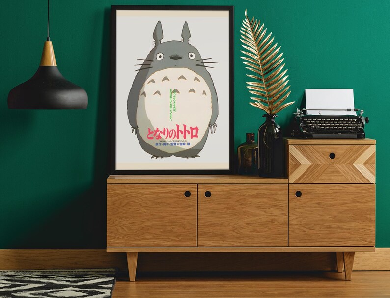 Pode incluir: Um poster emoldurado com o personagem Totoro do filme de anima&ccedil;&atilde;o Meu Vizinho Totoro. O poster &eacute; exibido em um arm&aacute;rio de madeira com uma m&aacute;quina de escrever preta, uma folha de ouro e uma lumin&aacute;ria pendente preta.