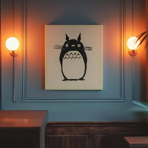 May include: A canvas print featuring a black and white illustration of Totoro from the animated film My Neighbor Totoro. The artwork is displayed on a blue wall, illuminated by warm-toned wall sconces. The print has a cream-colored background.