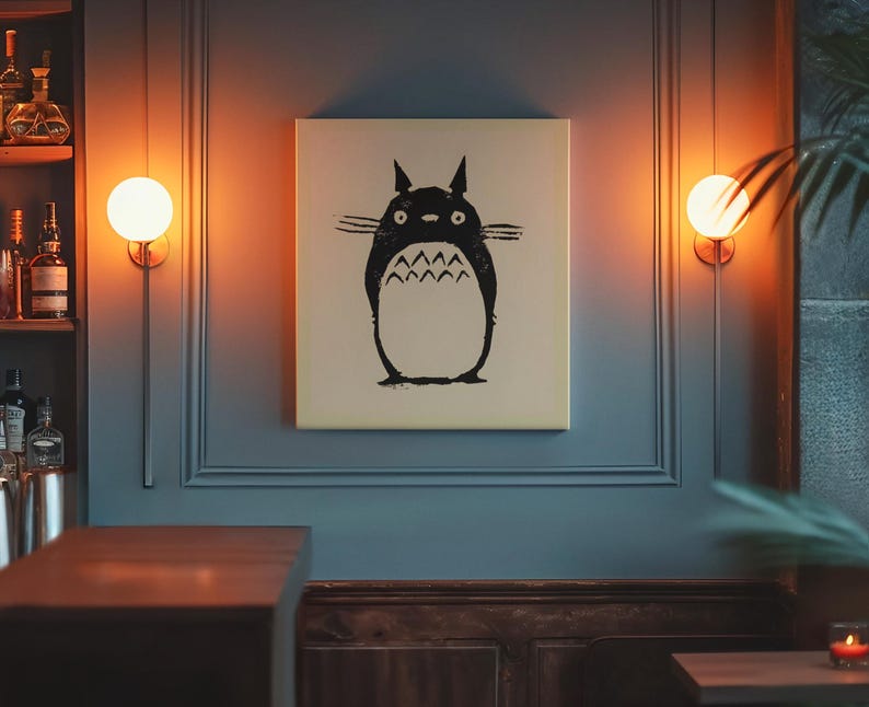 May include: A canvas print featuring a black and white illustration of Totoro from the animated film My Neighbor Totoro. The artwork is displayed on a blue wall, illuminated by warm-toned wall sconces. The print has a cream-colored background.