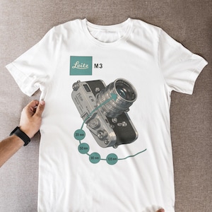Può includere: T-shirt bianca con un'illustrazione dettagliata di una fotocamera Leica M3 e del suo obiettivo. Il design include il testo "Leitz M3" in teal, insieme alle misure di 35 mm, 50 mm, 90 mm e 135 mm. La maglietta è tenuta da due mani.