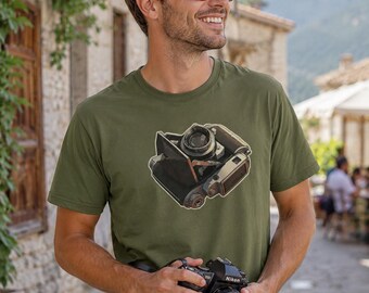 May include: An olive green short-sleeve t-shirt featuring a graphic illustration of a vintage folding camera in the center. The camera graphic displays black and metallic silver details with a lens and bellows mechanism visible in a stylized design.