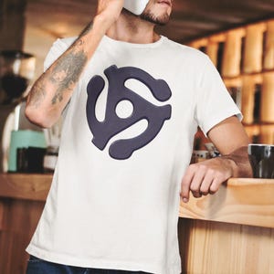 May include: White t-shirt featuring a dark blue 45 RPM record adapter graphic. The shirt is a classic crew neck style, perfect for music lovers. The design is centered on the chest.