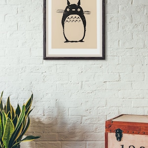 May include: A framed print featuring a black and white illustration of a Totoro character. The artwork is set against a beige background and framed in a dark brown frame. The print is mounted on a white brick wall.
