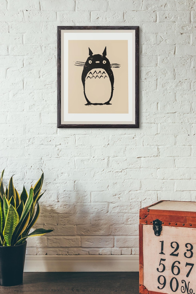May include: A framed print featuring a black and white illustration of a Totoro character. The artwork is set against a beige background and framed in a dark brown frame. The print is mounted on a white brick wall.