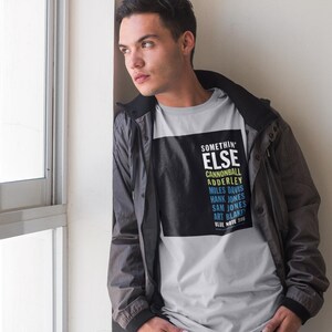 May include: A light grey t-shirt featuring the text "SOMETHIN' ELSE" and the names of jazz musicians, including Cannonball Adderley and Miles Davis. The person is wearing a grey and black jacket.