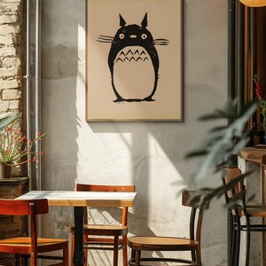 May include: A framed print featuring a black illustration of a Totoro character. The artwork is displayed on a neutral-toned background, mounted in a simple wooden frame. The image is set in a cafe with wooden chairs and a small table.