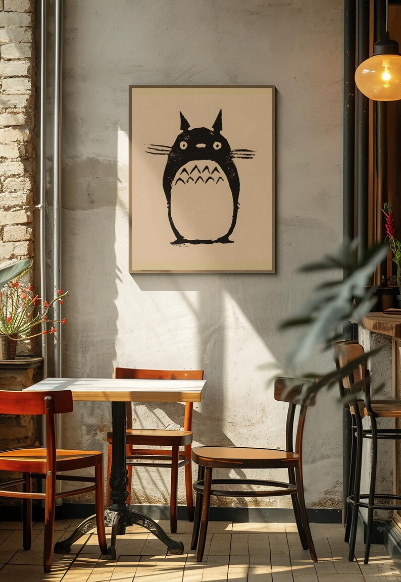 May include: A framed print featuring a black illustration of a Totoro character. The artwork is displayed on a neutral-toned background, mounted in a simple wooden frame. The image is set in a cafe with wooden chairs and a small table.