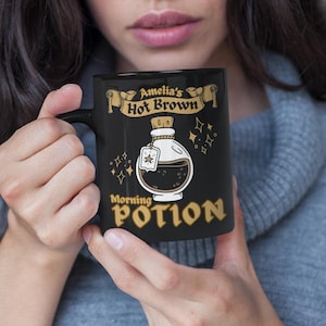 May include: Black coffee mug with a gold design featuring a potion bottle and the text "Amelia's Hot Brown Morning Potion".