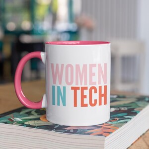 Puede incluir: Taza de cerámica blanca con borde y asa de color rosa. La taza tiene el texto "WOMEN IN TECH" impreso en rosa, azul y naranja.