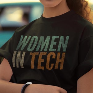 May include: A woman wearing a black t-shirt with the text "WOMEN IN TECH" in green, orange, and white. She is standing in front of a teal car and has her hair pulled back in a bun.