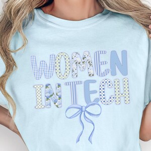 Può includere: T-shirt azzurra con la scritta "WOMEN IN TECH" in una miscela di caratteri fantasia e un fiocco grafico. La maglietta è realizzata in un materiale morbido e ha uno scollo tondo.