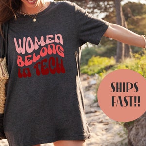 Puede incluir: Camiseta gris jaspeado oscuro con el texto "WOMEN BELONG IN TECH" en rosa y rojo. La camiseta es de cuello redondo y parece estar hecha de un material suave. Las palabras "DARK HEATHER GREY" están en la parte inferior de la imagen.