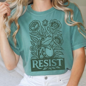 May include: A teal green t-shirt with a detailed illustration of a frog surrounded by flowers and foliage. The word "RESIST" is printed in a rectangular box at the bottom of the design. The shirt is made of a soft material.