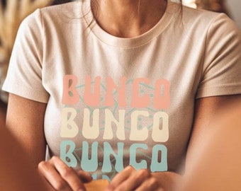 Puede incluir: Una camiseta beige con la palabra "BUNCO" apilada tres veces en colores pastel. La camiseta la lleva una persona con el pelo castaño recogido en un moño desordenado. La persona sonríe y parece estar en un café o restaurante.