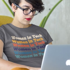 Puede incluir: Una persona con una camiseta gris con el texto "Women in Tech" repetido en colores arcoíris, sentada en una silla amarilla y usando un portátil plateado.