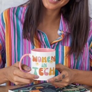 Peut inclure: Une tasse en céramique rose et blanche avec le texte "Women in Tech" en lettres colorées de style rétro. La tasse est décorée de petits accents de fleurs.