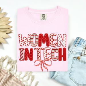 May include: A light pink t-shirt with the words "WOMEN IN TECH" in red, patterned letters. The letters have polka dots, stripes, and strawberry designs. A pink bow is below the text. The shirt is folded on a white surface.