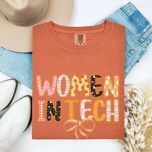 May include: An orange Comfort Colors t-shirt with the words "WOMEN IN TECH" in a colourful, patterned font. The design includes pink, white, orange, and black elements, with a bow at the bottom. The shirt is laid flat with a hat, jeans, and boots.