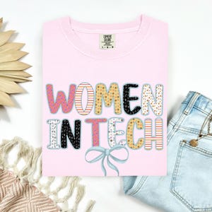 Könnte beinhalten: Hellrosa T-Shirt mit dem Text "WOMEN IN TECH" in bunten, gemusterten Buchstaben. Unter dem Text befindet sich eine hellblaue Schleife. Das Shirt liegt gefaltet auf einer weißen Oberfläche, mit einer Jeanshose.