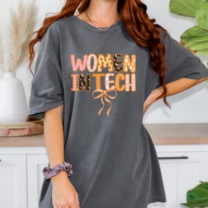 May include: Dark grey t-shirt with the text "WOMEN IN TECH" in a colourful, patterned design. The shirt features a bow graphic below the text. The shirt is a comfortable, casual style.