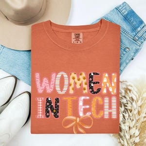 May include: An orange Comfort Colors t-shirt with the text "WOMEN IN TECH" in a colourful, patterned design. The shirt features a bow graphic. A tan hat, blue jeans, and white boots are visible.