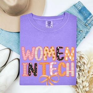 May include: A lilac Comfort Colors t-shirt with the words "WOMEN IN TECH" in a decorative font. The letters are decorated with patterns and Halloween-themed graphics, including pumpkins and ghosts. A bow graphic is at the bottom.