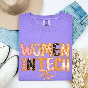 May include: A lilac t-shirt featuring the text "WOMEN IN TECH" in a colourful, patterned design. The design includes pumpkins, ghosts, and a bow. The shirt is a Comfort Colors brand.