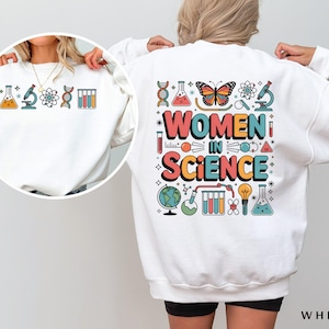 Puede incluir: Sudadera blanca con un diseño gráfico colorido que incluye el texto "Women in Science" y varios iconos relacionados con la ciencia, como una mariposa, un microscopio, un vaso de precipitados y una cadena de ADN.