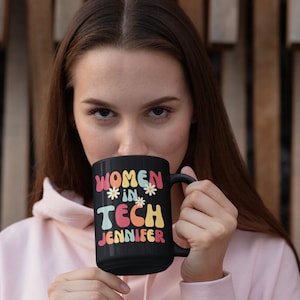 Puede incluir: Taza de café negra con el texto "Women in Tech" en letras de colores y el nombre "Jennifer" debajo. La taza tiene un diseño de margaritas blancas.