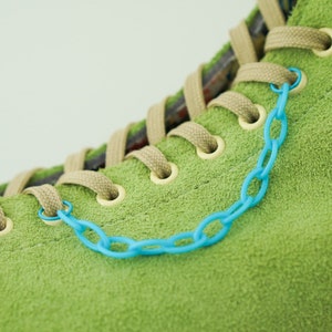 Op de afbeelding: Een groene suède schoen met bruine veters en een detail van een blauwe plastic ketting.