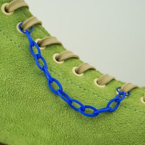 May include: Green suede shoe with a blue plastic chain detail. The shoe has beige laces and a white sole.