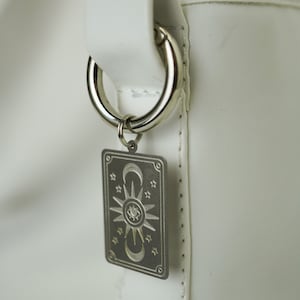 May include: White leather tote bag with a silver ring and a silver charm featuring a sun, moon, and eye design.