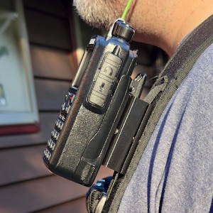 May include: A black two-way radio with a microphone attached to a black shoulder strap. The radio is clipped to the strap with a black clip.