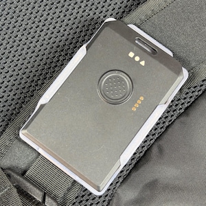 May include: A black rectangular device with a circular button and indicator lights, attached to a black backpack strap. The device has a white border and is made of plastic. The background is a black mesh material.