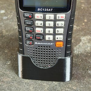 May include: A Uniden BC125AT handheld scanner with a black and silver design. The scanner has a small screen, a keypad with various function buttons, and an antenna. It is placed in a black holder.