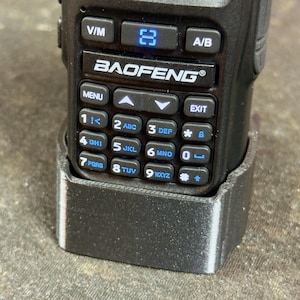 May include: A black Baofeng radio with a digital display showing frequency readings. The radio has a keypad with numbered buttons and function keys. The display shows the frequency 146.960 and 447.000. The radio is in a black holder.