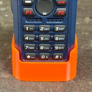May include: A blue handheld radio with an orange charging base. The radio has a keypad with numbers and function buttons. The screen displays "HS PNWR 2" and "PNW Rgnl 2".