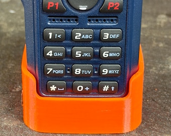 Pode incluir: Um rádio portátil azul com uma base de carregamento laranja. O rádio tem um teclado com números e botões de função. O ecrã mostra "HS PNWR 2" e "PNW Rgnl 2".