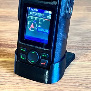 May include: A black handheld two-way radio with a blue screen displaying "OLYCOMM3" and "WWRNQ66". The radio has a black rubberized case and a black stand.