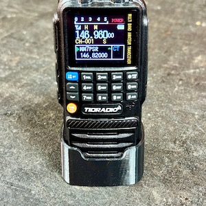 May include: A black Tidradio multi-band amateur transceiver radio with a digital display showing frequency information. The radio is in a black holder. The display shows the frequency 146.96000.