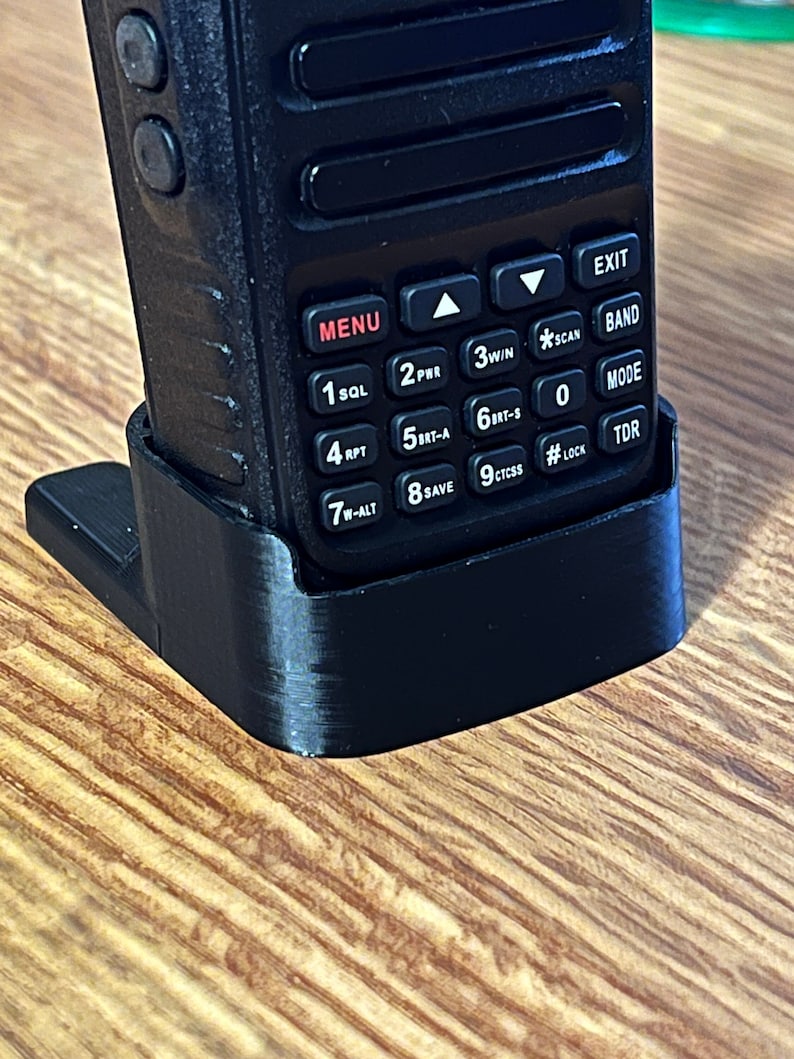 Pode incluir: Um r&aacute;dio port&aacute;til preto est&aacute; em um suporte preto. O r&aacute;dio tem um teclado com bot&otilde;es rotulados MENU, EXIT, SQL, PHR, WIN, SCAN, RPT, Brt-A, Brt-S, ALT, SAVE, CTCSS, LOCK e TDR. O suporte est&aacute; em uma superf&iacute;cie de madeira.