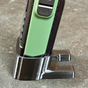 May include: A black 3D-printed cradle supporting a black and green handheld device. The cradle features a rectangular base with two supporting arms. The handheld device has a green side panel and a microphone icon.